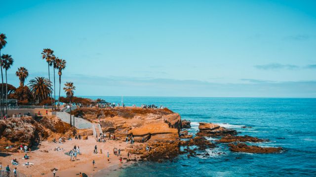 A sunny rocky coastline with palm trees, waves crashing, and people relaxing on the sandy beach.