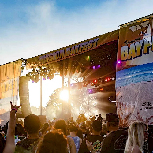Crowd watches a live concert on the “Mission Bayfest” outdoor stage as colorful lights flare in the sunset.