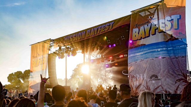 Crowd watches a live concert on the &ldquo;Mission Bayfest&rdquo; outdoor stage as colorful lights flare in the sunset.