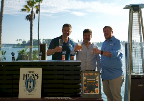 Three men smile and toast with beer glasses at an outdoor lakeside event with palm trees, beside a beer tap dispenser.