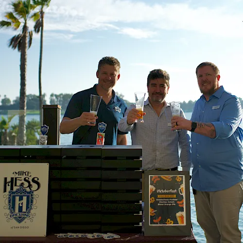 Three men smile and toast with beer glasses at an outdoor lakeside event with palm trees, beside a beer tap dispenser.