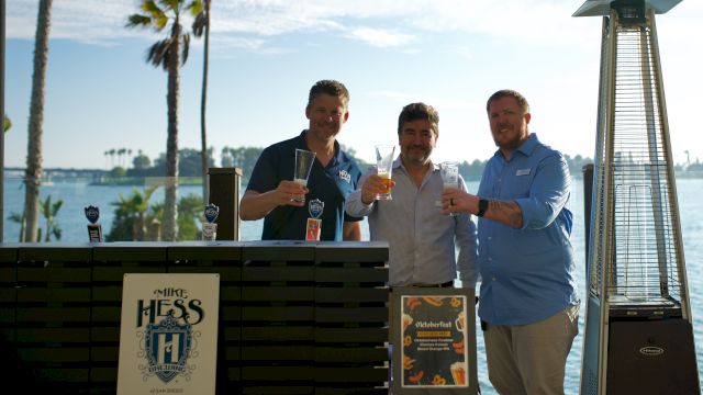 Three men smile and toast with beer glasses at an outdoor lakeside event with palm trees, beside a beer tap dispenser.