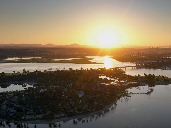 Golden sunset over a wide river with a long bridge, islands, and distant hills in a calm aerial view.