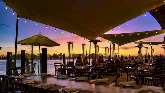 A waterfront dining patio at sunset with string lights, tables, chairs, and tiki torches glowing along the river.