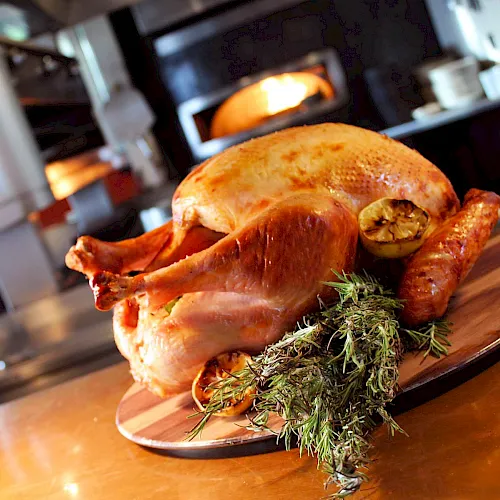 A roasted chicken sits on a wooden board, topped with fresh herbs, in front of a lit oven.