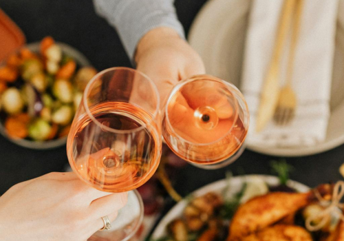 Two people clink pink wine glasses over a table of roasted food and salad.