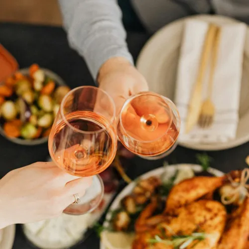 Two people clink pink wine glasses over a table of roasted food and salad.