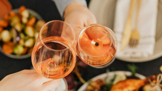 Two people clink pink wine glasses over a table of roasted food and salad.