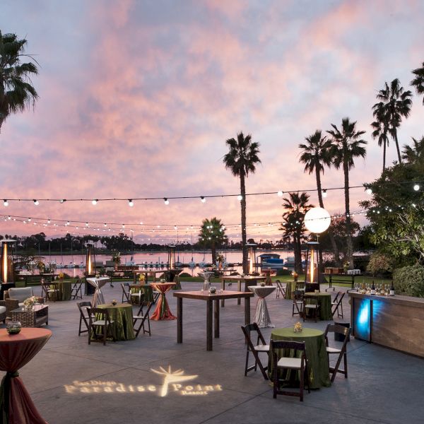 An outdoor event patio at sunset with palm trees, string lights, decorated tables, and a glowing pool backdrop.