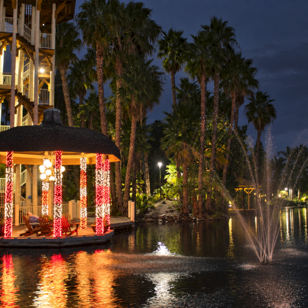 A lit, red-decorated gazebo glows on a lakeside at dusk as palm trees and fireworks light the night.