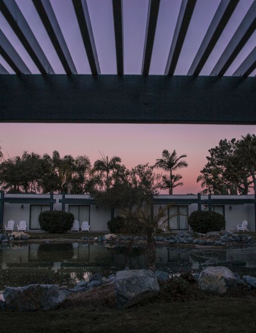 A tropical resort scene at dusk: palm trees, a pond/pool reflection, and white bungalows under a purple-pink sky.
