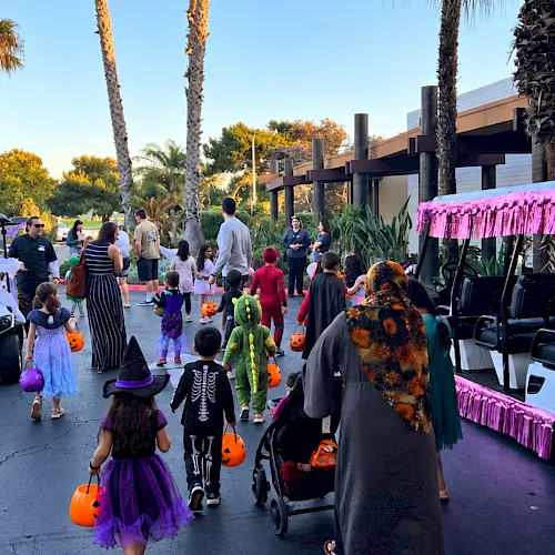 A Halloween trick-or-treat parade with kids in costumes, walking between decorated golf carts and a palm-lined outdoor venue.
