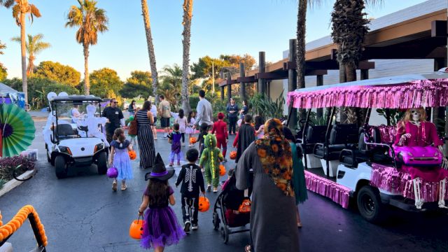 A Halloween trick-or-treat parade with kids in costumes, walking between decorated golf carts and a palm-lined outdoor venue.
