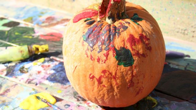 A painted orange pumpkin covered in colorful red, blue, green, and purple paint on a messy art-station table.