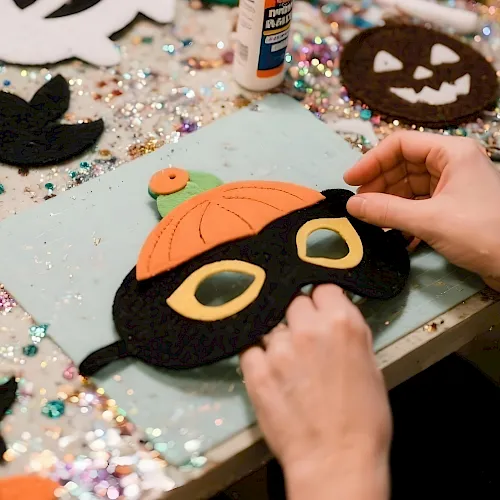 A person&rsquo;s hands craft Halloween felt with a jack-o&rsquo;-lantern face on a glittery table covered in decorations.