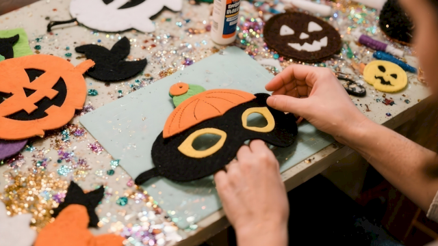 A person&rsquo;s hands craft Halloween felt with a jack-o&rsquo;-lantern face on a glittery table covered in decorations.